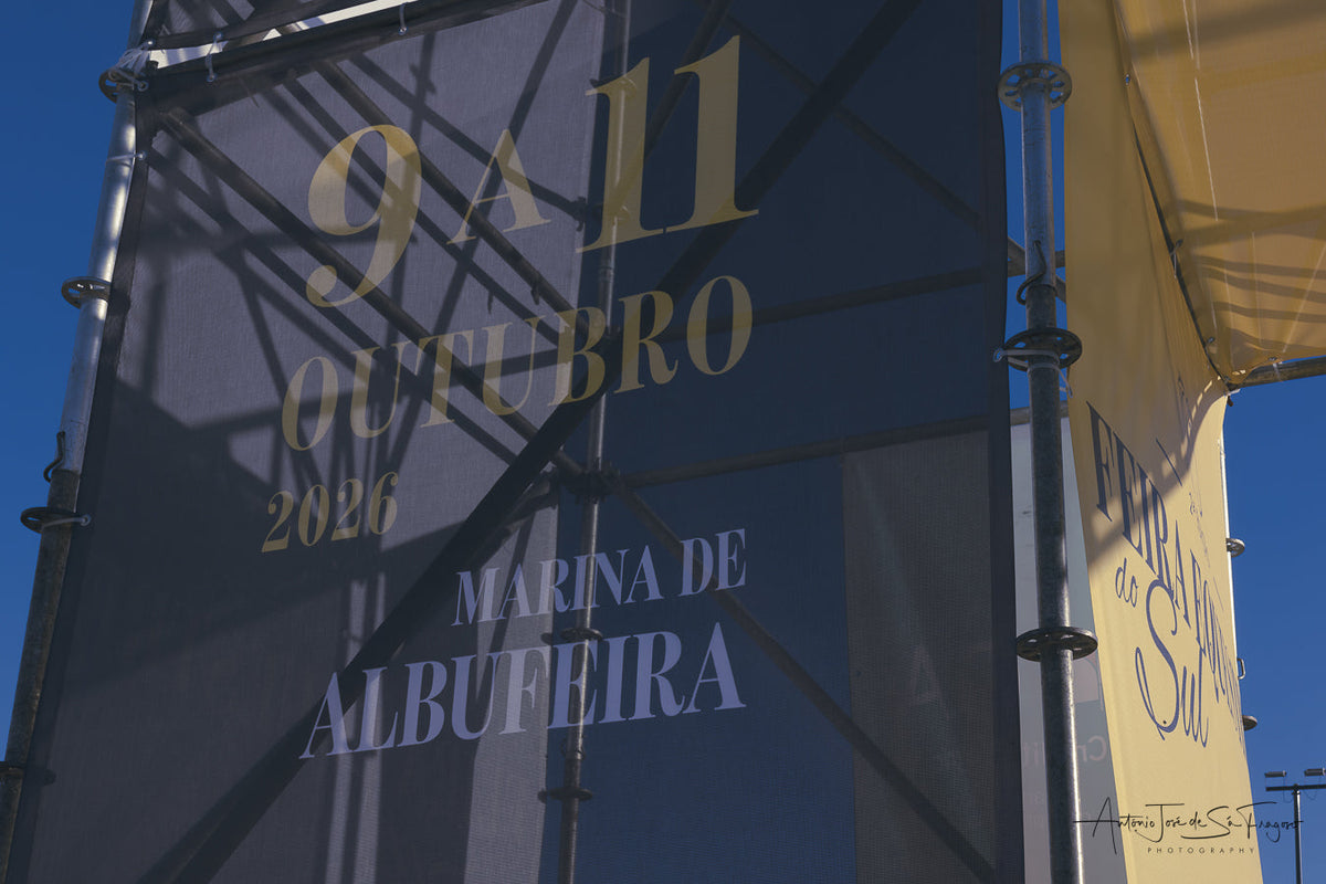 A close-up of a large dark blue banner printed with the text '9 A 11 OUTUBRO 2026 MARINA DE ALBUFEIRA' in gold and white lettering, attached to a metal support structure against a blue sky.