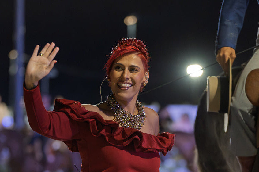 A woman in a red ruffled top and black skirt waves to a crowd at night while standing next to a horse, with stadium lights blurred in the background.