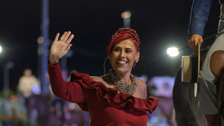 A woman in a red ruffled top and black skirt waves to a crowd at night while standing next to a horse, with stadium lights blurred in the background.