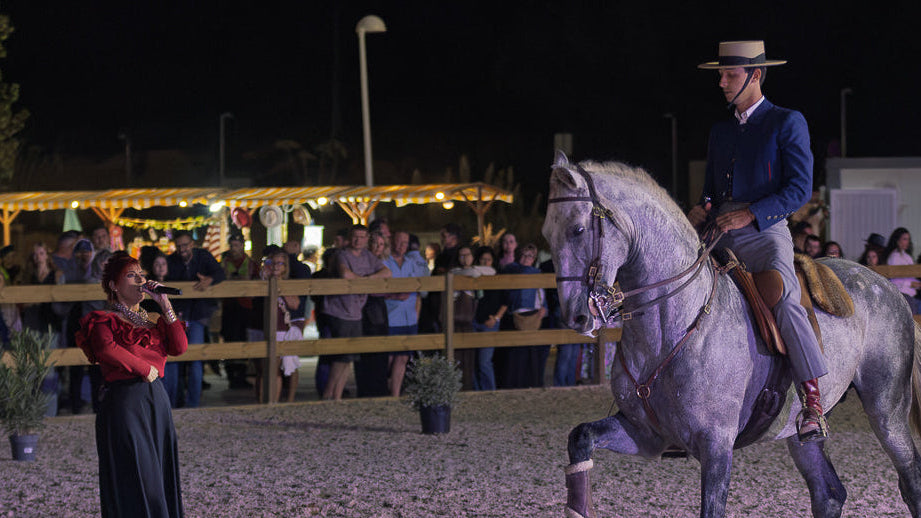 A man in a portugueses traditional hat rides a dappled gray horse during an evening event, with a crowd of spectators visible behind a wooden fence in the background.