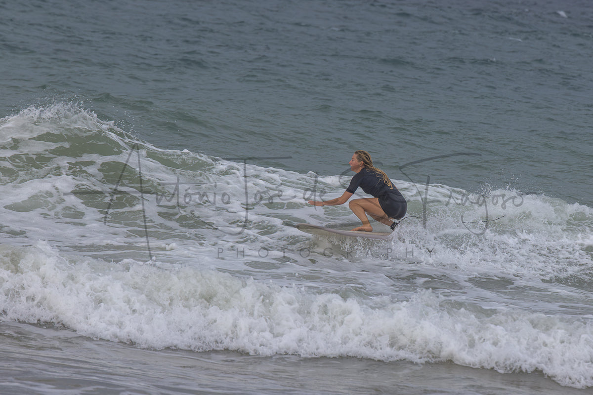 A person in a dark swimsuit crouches on a surfboard on a breaking ocean wave, with white sea foam and gray-blue water around them. A large black script watermark reading 'Antonio José de S.