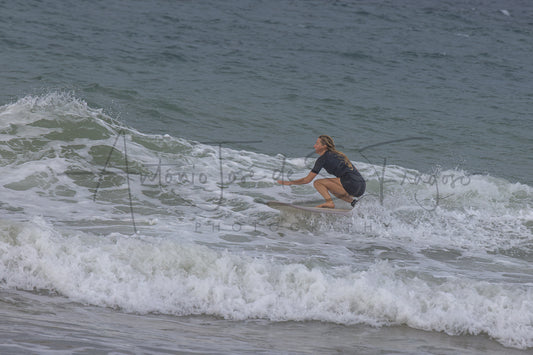 A person in a dark swimsuit crouches on a surfboard on a breaking ocean wave, with white sea foam and gray-blue water around them. A large black script watermark reading 'Antonio José de S.