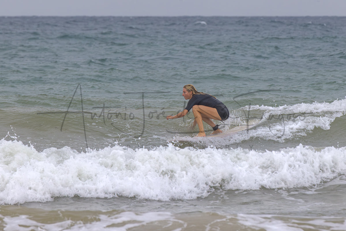 A surfer crouches on a white surfboard in the ocean waves, wearing a dark gray short-sleeve top and black shorts. A large cursive watermark is visible across the center of the image.