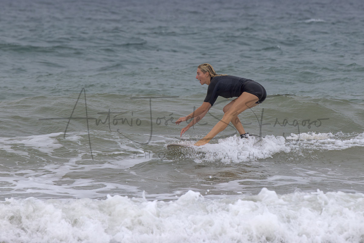 A woman in a black swimsuit stands on a surfboard in the ocean waves, with white foam in the foreground and a watermark across the center.