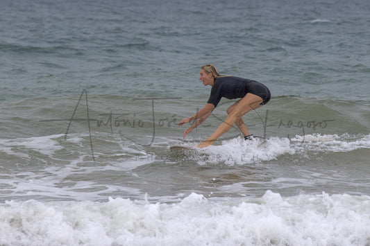 A woman in a black swimsuit stands on a surfboard in the ocean waves, with white foam in the foreground and a watermark across the center.