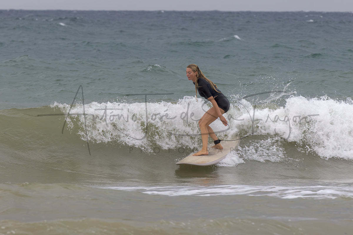 A woman in a black swimsuit stands on a light-colored surfboard riding a breaking ocean wave, with the sea and cloudy sky in the background.