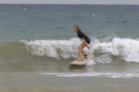 A woman in a black swimsuit stands on a light-colored surfboard riding a breaking ocean wave, with the sea and cloudy sky in the background.