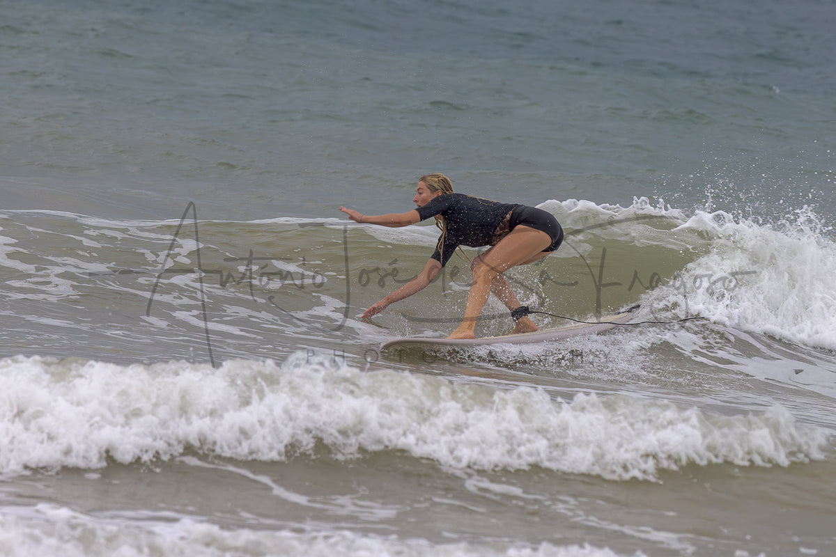 A woman in a black swimsuit rides a surfboard on a small ocean wave, with white foam and gray-blue water around her.