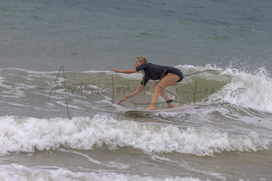 A woman in a black swimsuit rides a surfboard on a small ocean wave, with white foam and gray-blue water around her.