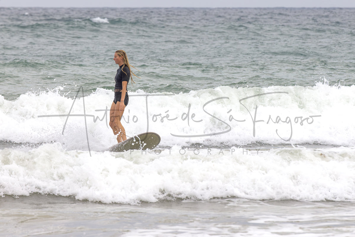 A woman in a black swimsuit stands on a surfboard in the ocean, riding a breaking wave with white foam around her. A large cursive photographer watermark is overlaid across the center.