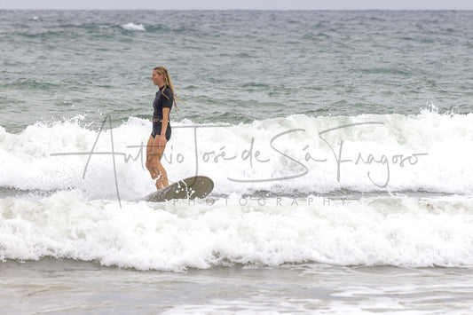 A woman in a black swimsuit stands on a surfboard in the ocean, riding a breaking wave with white foam around her. A large cursive photographer watermark is overlaid across the center.