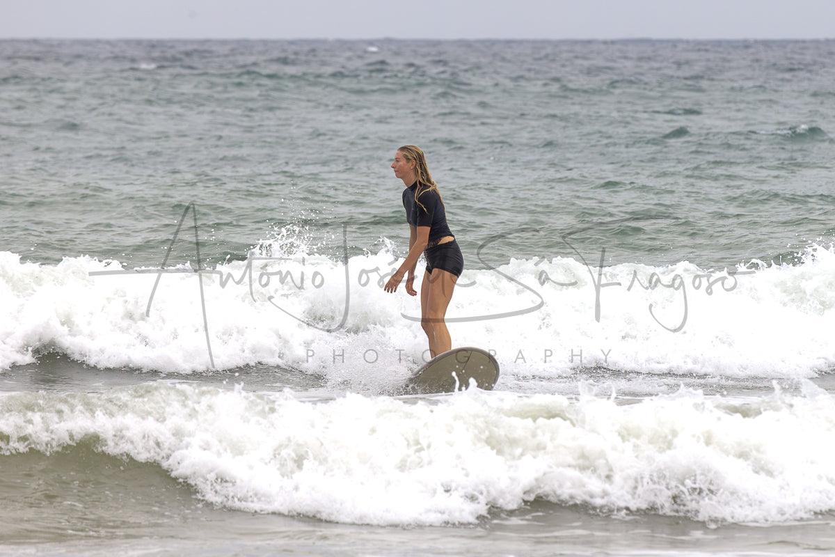 A woman in a black swimsuit stands in foamy ocean waves under an overcast sky, with a large 'Antonio Jose Sargadelos Photography' watermark across the center.