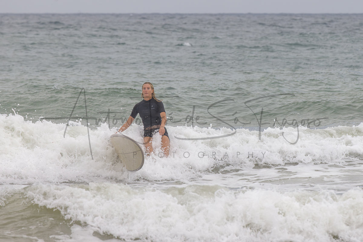 A surfer wearing a black wetsuit rides a white surfboard through white ocean waves under an overcast sky.
