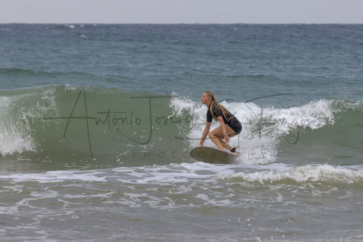 A woman in a black and yellow wetsuit rides a surfboard on a breaking ocean wave, with the ocean and sky in the background and a visible photographer watermark across the center.