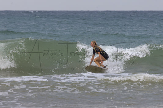 A woman in a black and yellow wetsuit rides a surfboard on a breaking ocean wave, with the ocean and sky in the background and a visible photographer watermark across the center.