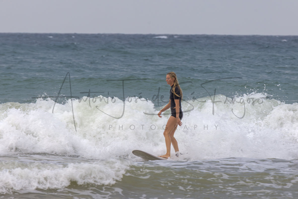 A woman in a black swimsuit stands on a surfboard in the surf of a wave, with the ocean and sky behind her. A large black script watermark reading 'Antonio José de Sá Fragoso' and 'PHOTOGRAPHY' is overlaid across the center.