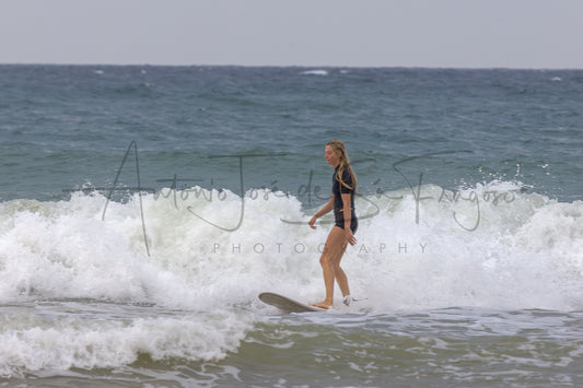 A woman in a black swimsuit stands on a surfboard in the surf of a wave, with the ocean and sky behind her. A large black script watermark reading 'Antonio José de Sá Fragoso' and 'PHOTOGRAPHY' is overlaid across the center.