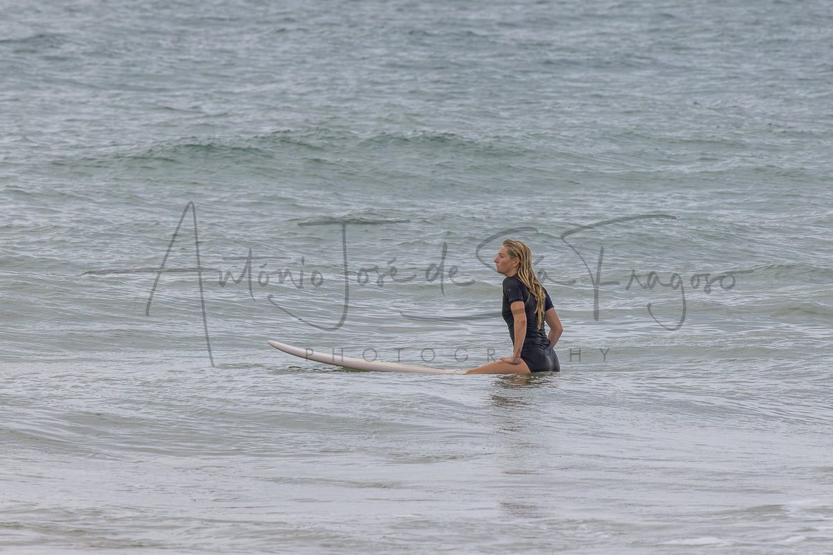 A woman in a black wetsuit sits on a white surfboard in shallow ocean water, with gentle waves around her. A large semi-transparent photographer watermark is visible across the center.