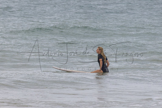 A woman in a black wetsuit sits on a white surfboard in shallow ocean water, with gentle waves around her. A large semi-transparent photographer watermark is visible across the center.