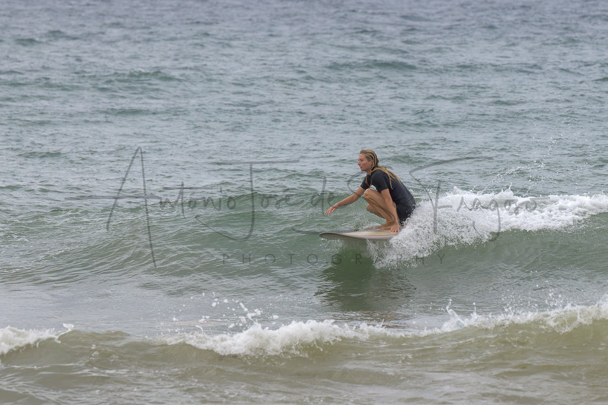 A surfer in a black wetsuit crouches on a surfboard riding a small ocean wave, with choppy gray-green water around them. A large semi-transparent photographer watermark is visible across the center.