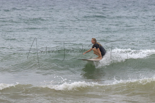 A surfer in a black wetsuit crouches on a surfboard riding a small ocean wave, with choppy gray-green water around them. A large semi-transparent photographer watermark is visible across the center.