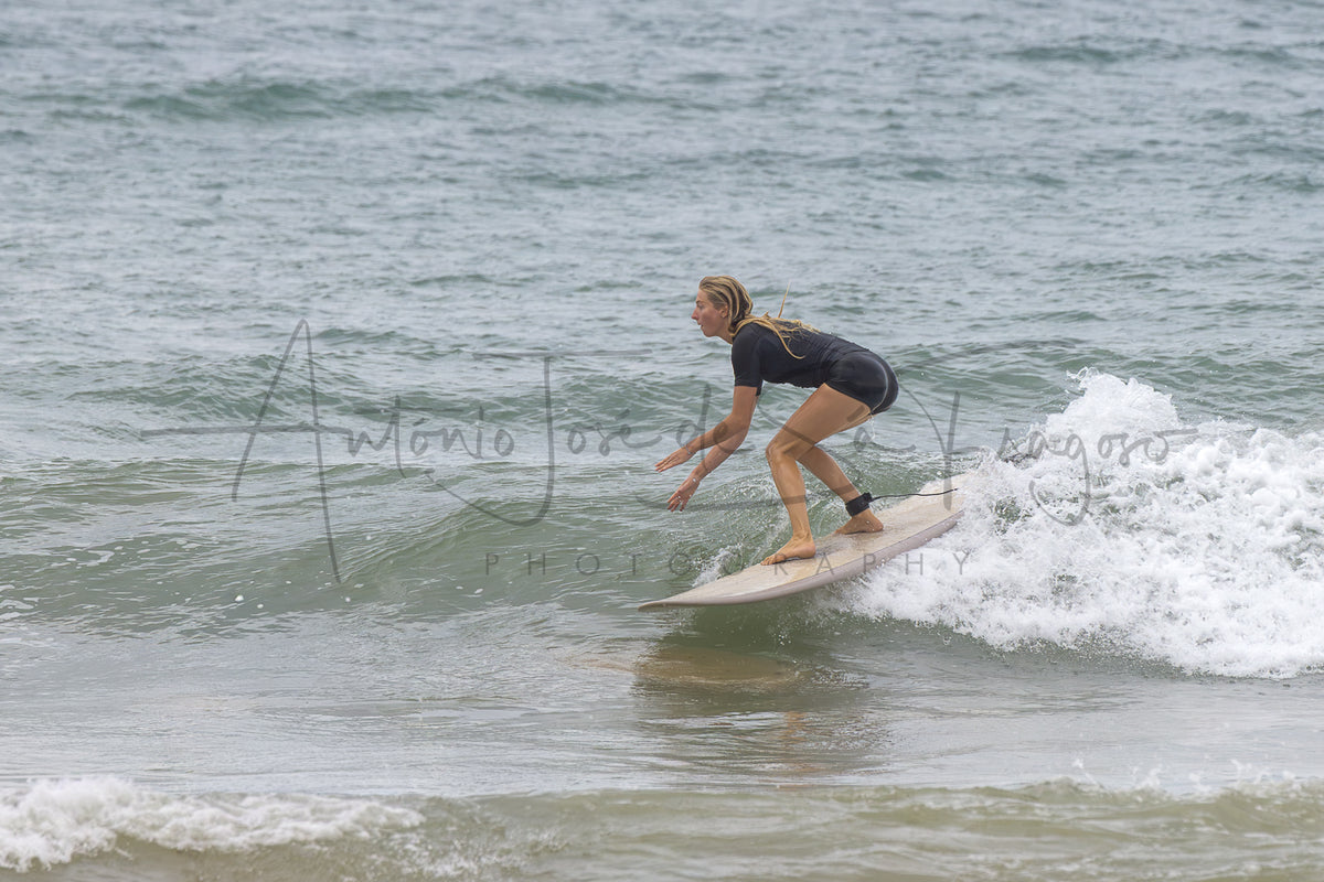 A surfer in a black wetsuit rides a wave on a white surfboard in the ocean, with a large photographer's watermark across the center.