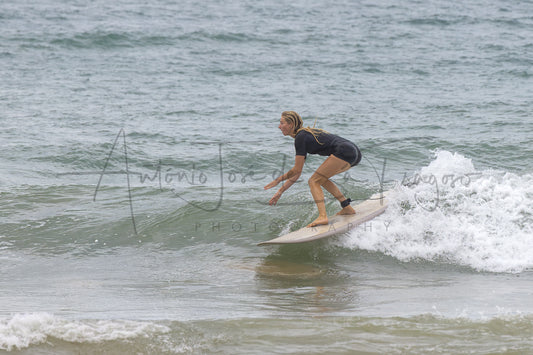 A surfer in a black wetsuit rides a wave on a white surfboard in the ocean, with a large photographer's watermark across the center.