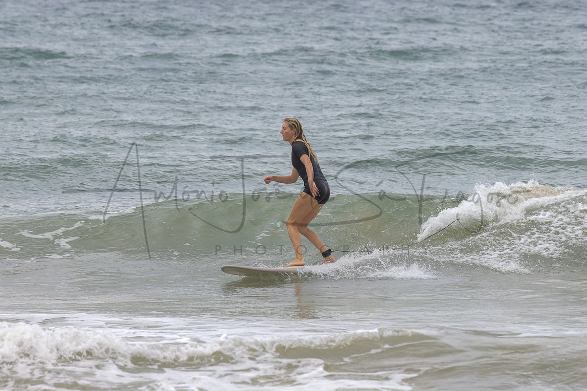 A woman in a black wetsuit stands on a surfboard in the ocean, riding a small wave with water splashing around her.