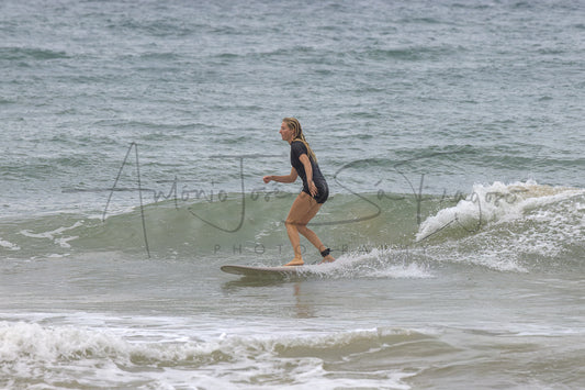 A woman in a black wetsuit stands on a surfboard in the ocean, riding a small wave with water splashing around her.