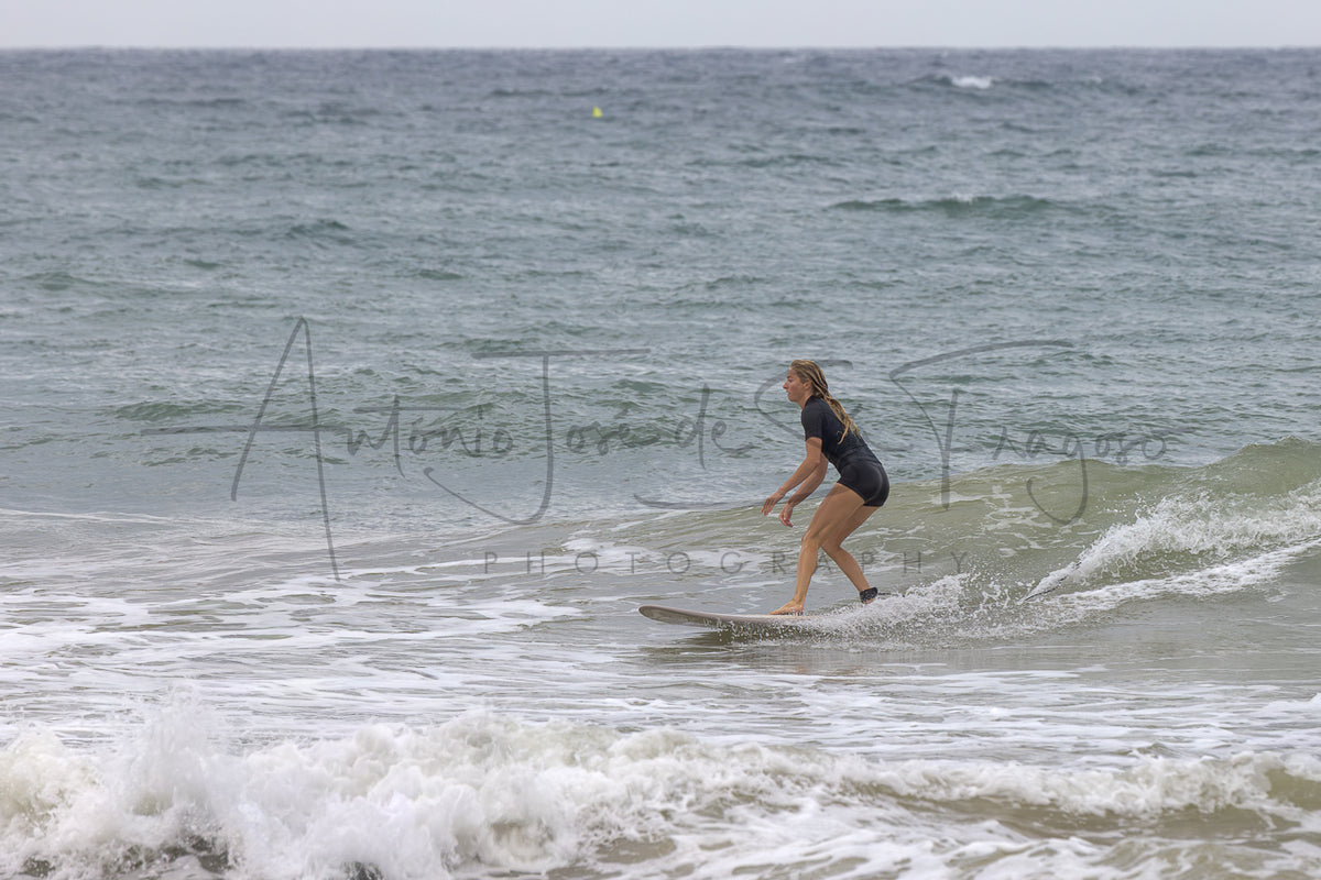 A woman in a black swimsuit stands on a surfboard in the ocean waves, with a large photographer watermark across the center.