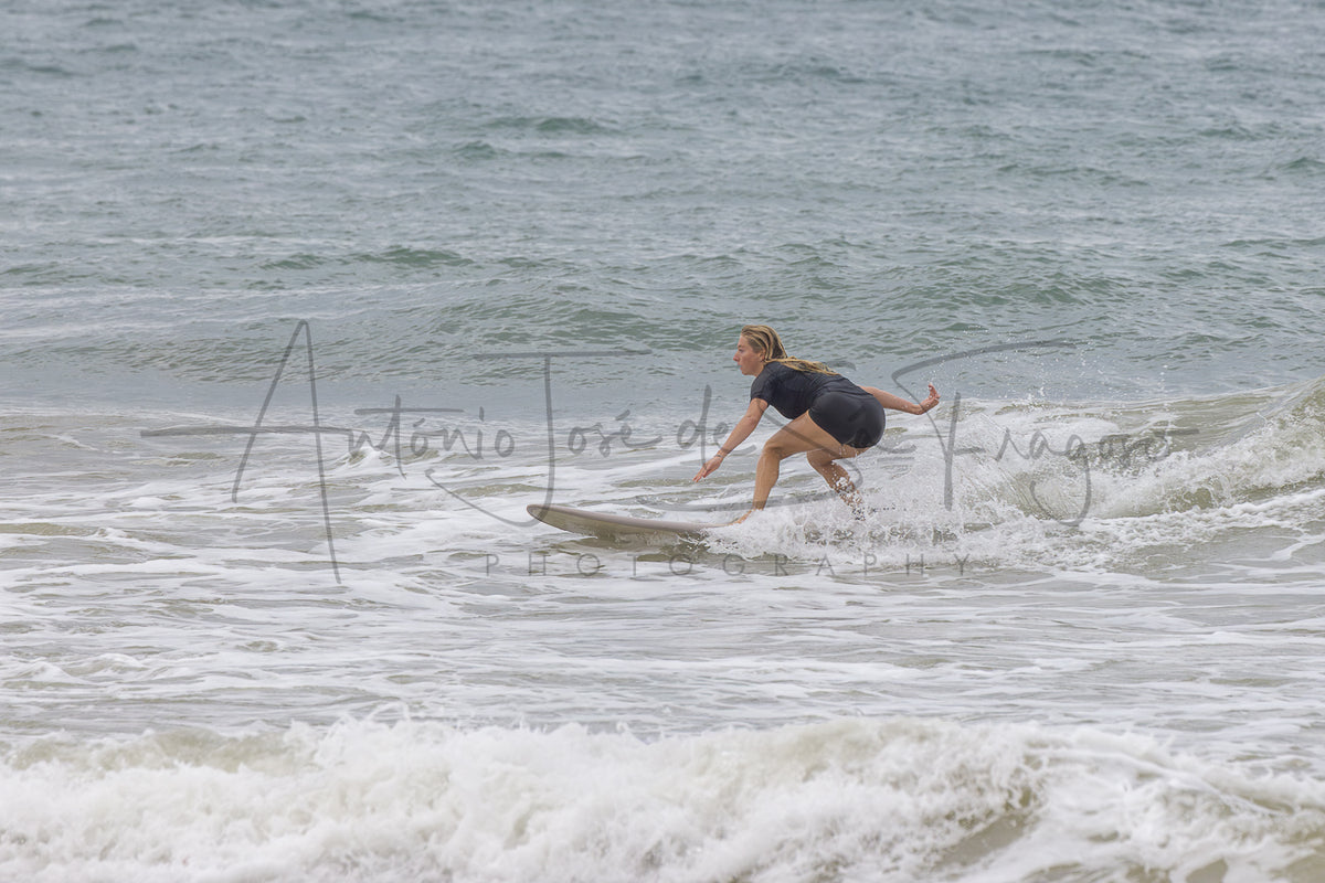A woman in a dark gray t-shirt and black shorts surfing on a wave, with a large watermark reading 'Antonio Jose de Sruiga Photography' across the center.