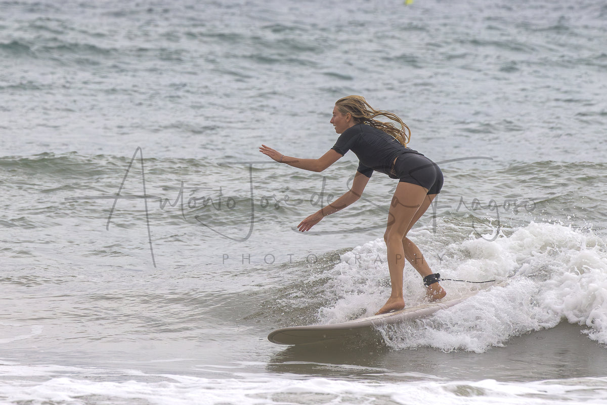 A woman in a black wetsuit stands on a surfboard on a small ocean wave, with ocean water and white foam around her. A large script watermark is visible across the center.