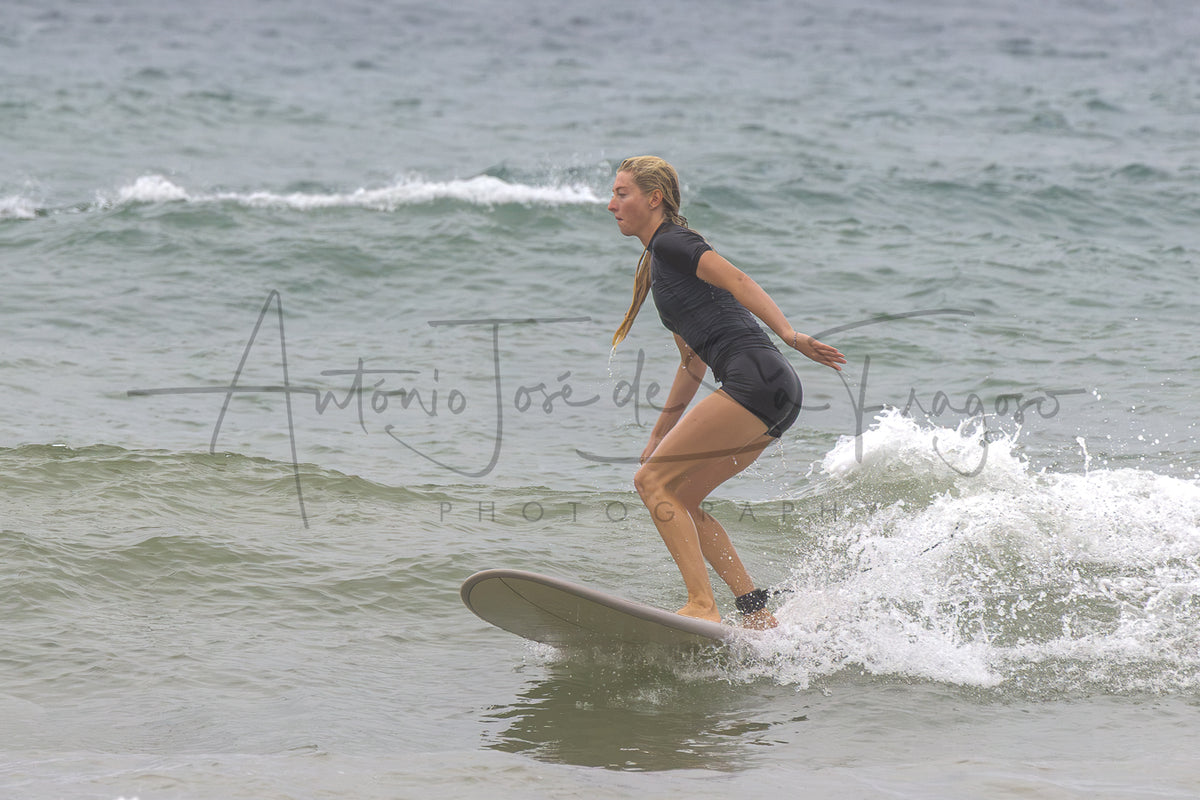 A woman in a black wetsuit stands on a surfboard in the ocean, balancing on a small wave. A large photographer's watermark is visible across the center of the image.
