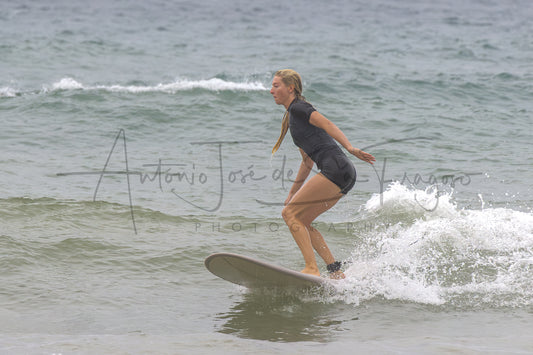 A woman in a black wetsuit stands on a surfboard in the ocean, balancing on a small wave. A large photographer's watermark is visible across the center of the image.