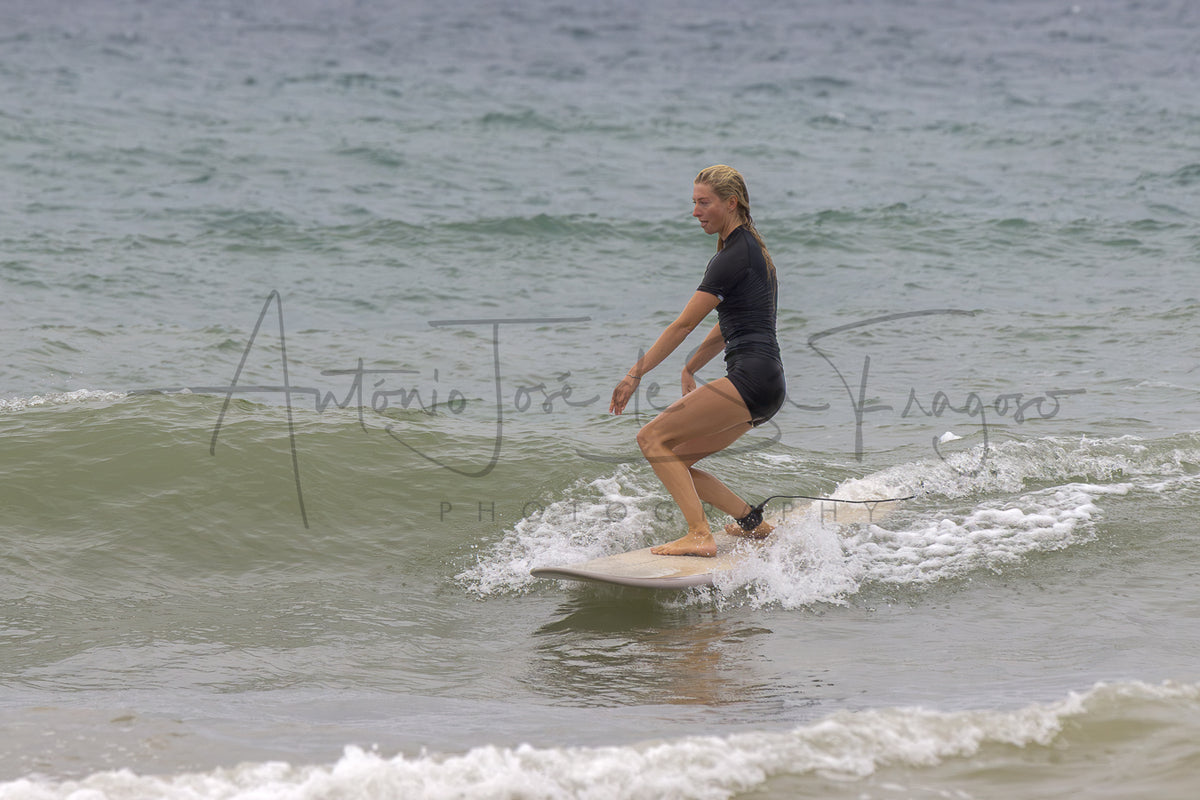 A woman in a black swimsuit stands on a white surfboard in the ocean, riding a small wave. A large photographer's watermark is overlaid across the center of the photo.