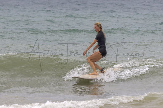 A woman in a black swimsuit stands on a white surfboard in the ocean, riding a small wave. A large photographer's watermark is overlaid across the center of the photo.