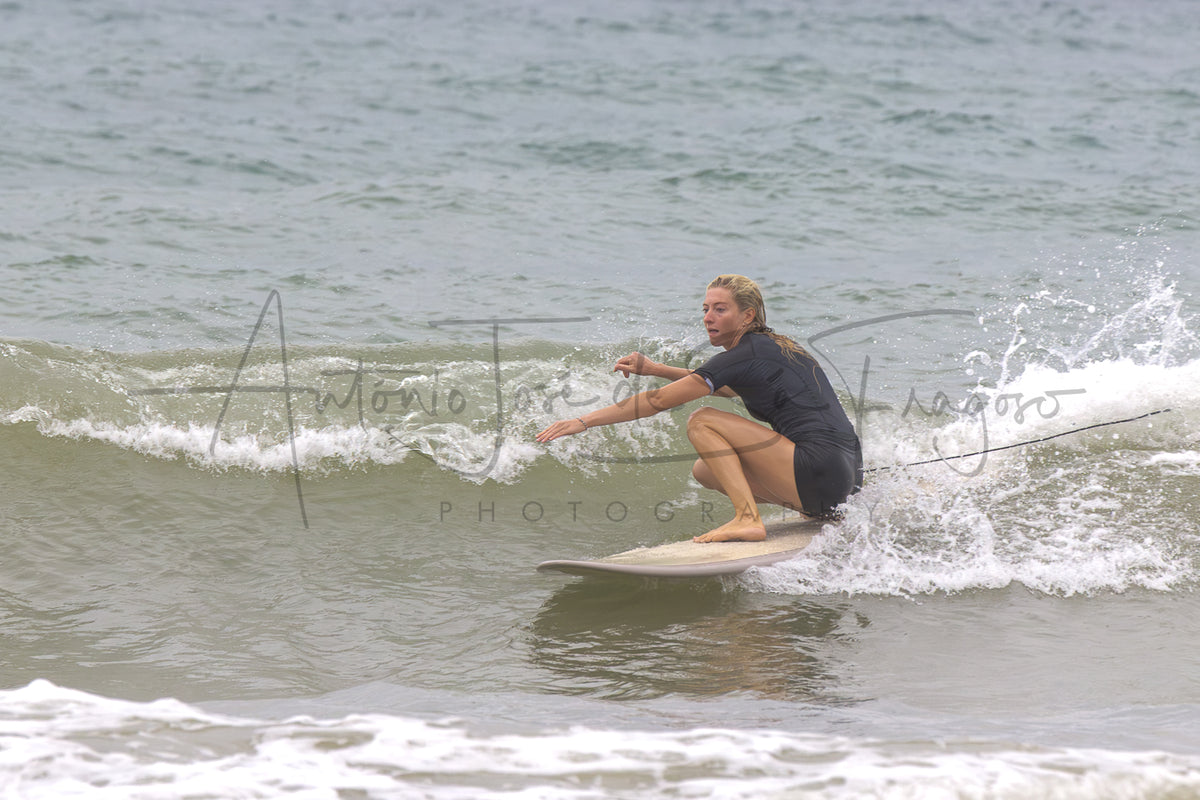 A surfer in a dark wetsuit crouches on a surfboard on a small ocean wave, with ocean water and white surf around them. A large photographer watermark is visible across the center.