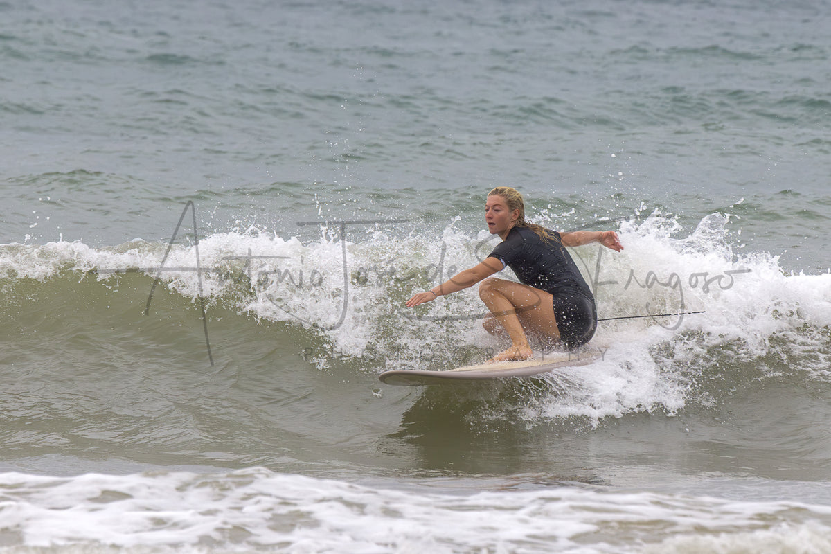 A surfer rides a breaking ocean wave, wearing a black wetsuit and crouching on a surfboard with white sea foam around them. The water is a muted gray-green, and a faint photographer watermark is visible across the wave.