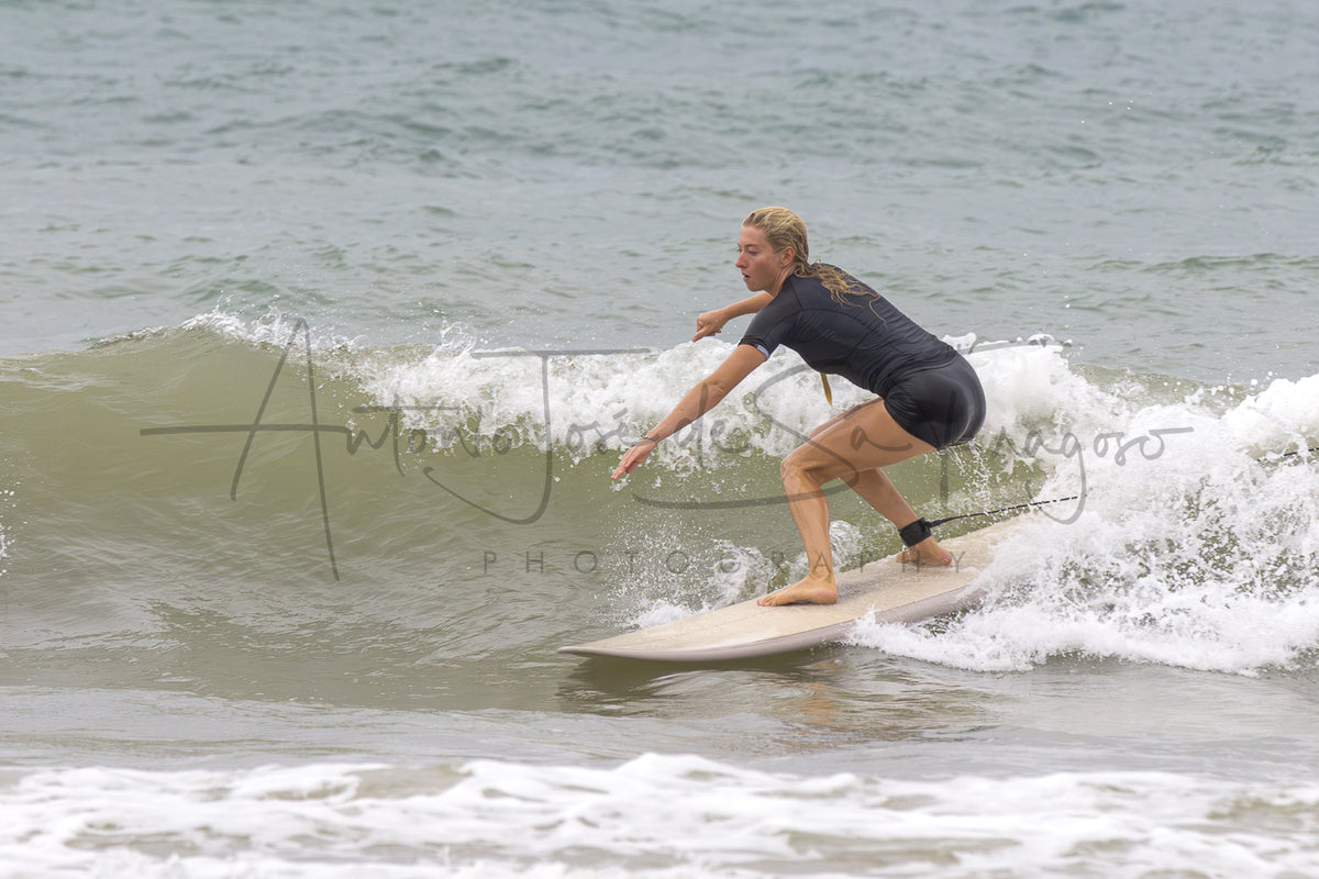 A woman in a black wetsuit stands on a light-colored surfboard, balancing on a breaking ocean wave. A large semi-transparent photographer watermark is visible across the center.