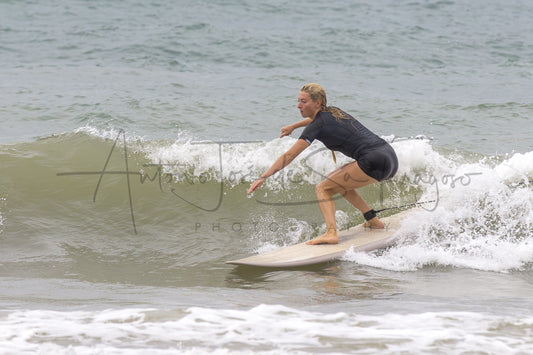A woman in a black wetsuit stands on a light-colored surfboard, balancing on a breaking ocean wave. A large semi-transparent photographer watermark is visible across the center.