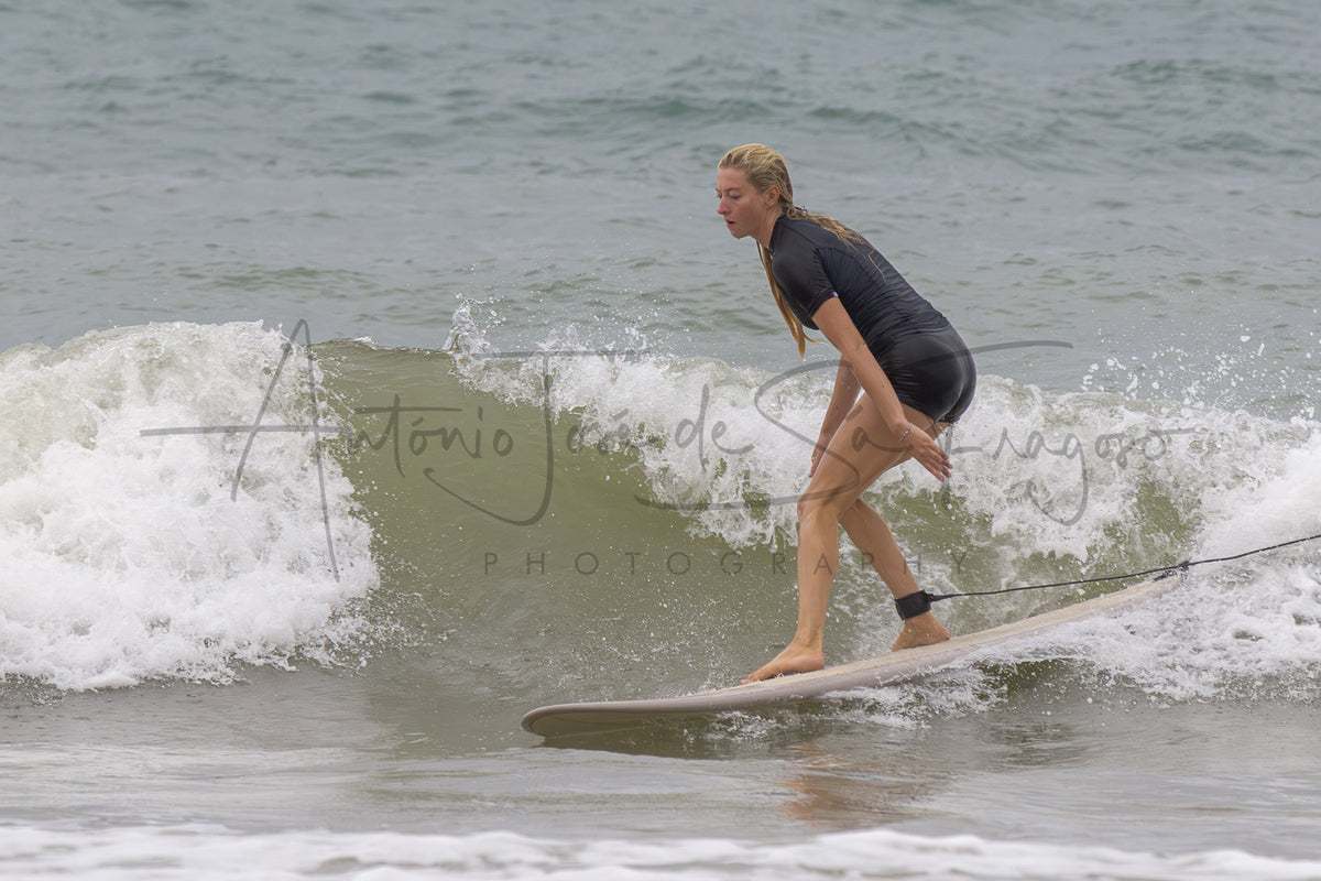 A woman in a black wetsuit stands on a surfboard in the ocean, riding a breaking wave. A large black script watermark reading 'Antonio José de Ságuro Photography' is overlaid across the center.