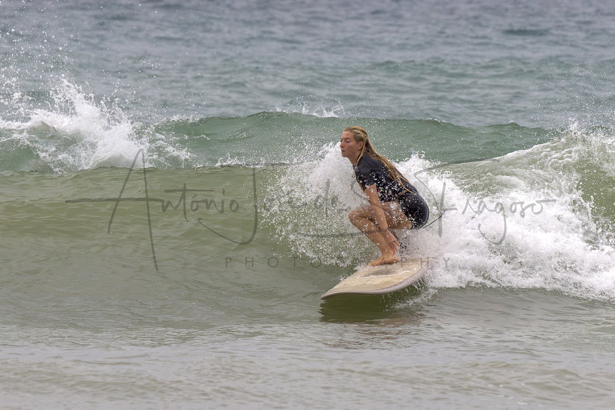 A surfer in a black swimsuit crouches on a light-colored surfboard riding a small ocean wave, with white spray and gray-blue water around them. A large photographer's watermark is visible across the center.