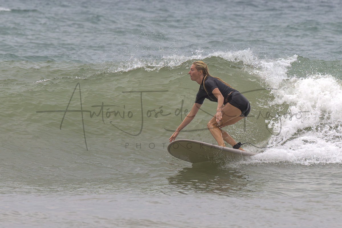 A woman in a black swimsuit rides a surfboard on a breaking ocean wave, with white sea foam and muted gray-green water in the background. A large photographer's watermark is visible across the center.