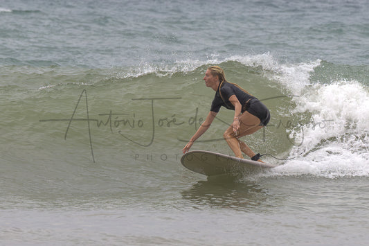 A woman in a black swimsuit rides a surfboard on a breaking ocean wave, with white sea foam and muted gray-green water in the background. A large photographer's watermark is visible across the center.