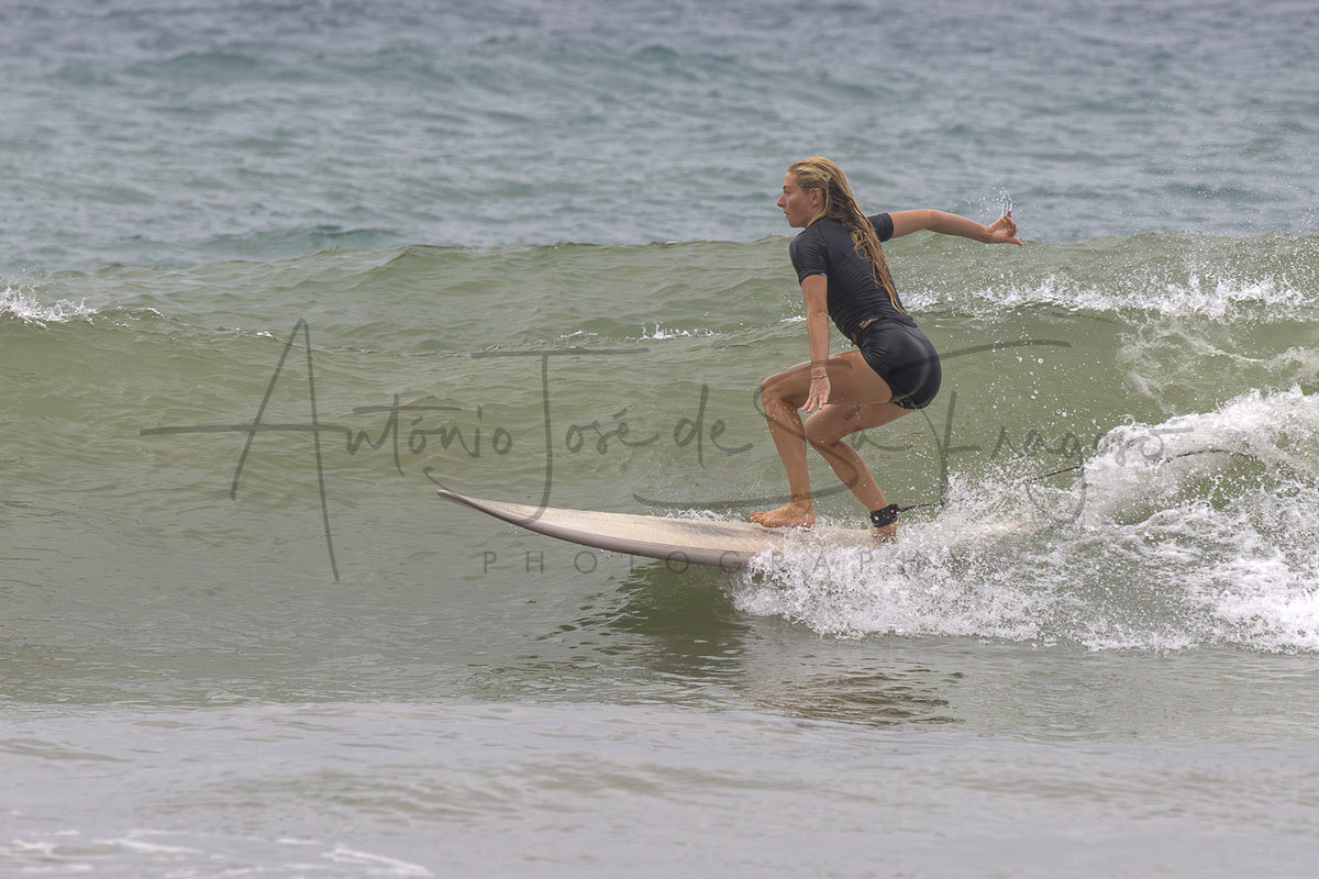 A woman in a black wetsuit rides a surfboard on a gentle ocean wave, with a large photographer's watermark across the center of the photo.