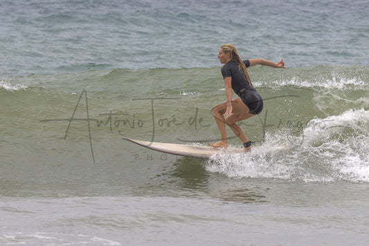 A woman in a black wetsuit rides a surfboard on a gentle ocean wave, with a large photographer's watermark across the center of the photo.