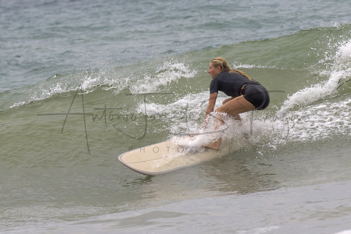 A woman in a black swimsuit crouches on a light-colored surfboard, riding a gentle ocean wave with white spray.