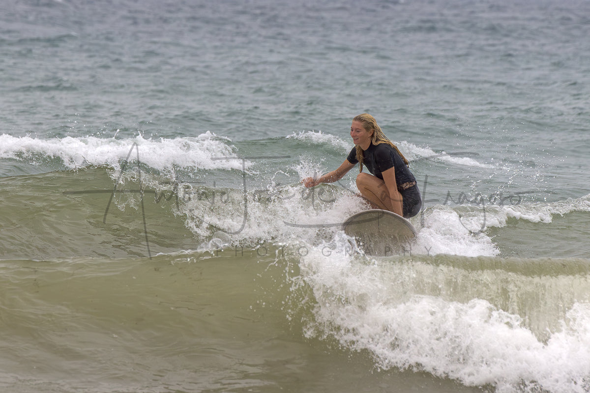 A woman in a black wetsuit crouches on a surfboard riding a small ocean wave, with white surf foam around her and a muted blue-gray sea in the background.