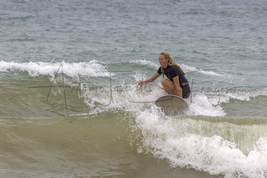 A woman in a black wetsuit crouches on a surfboard riding a small ocean wave, with white surf foam around her and a muted blue-gray sea in the background.