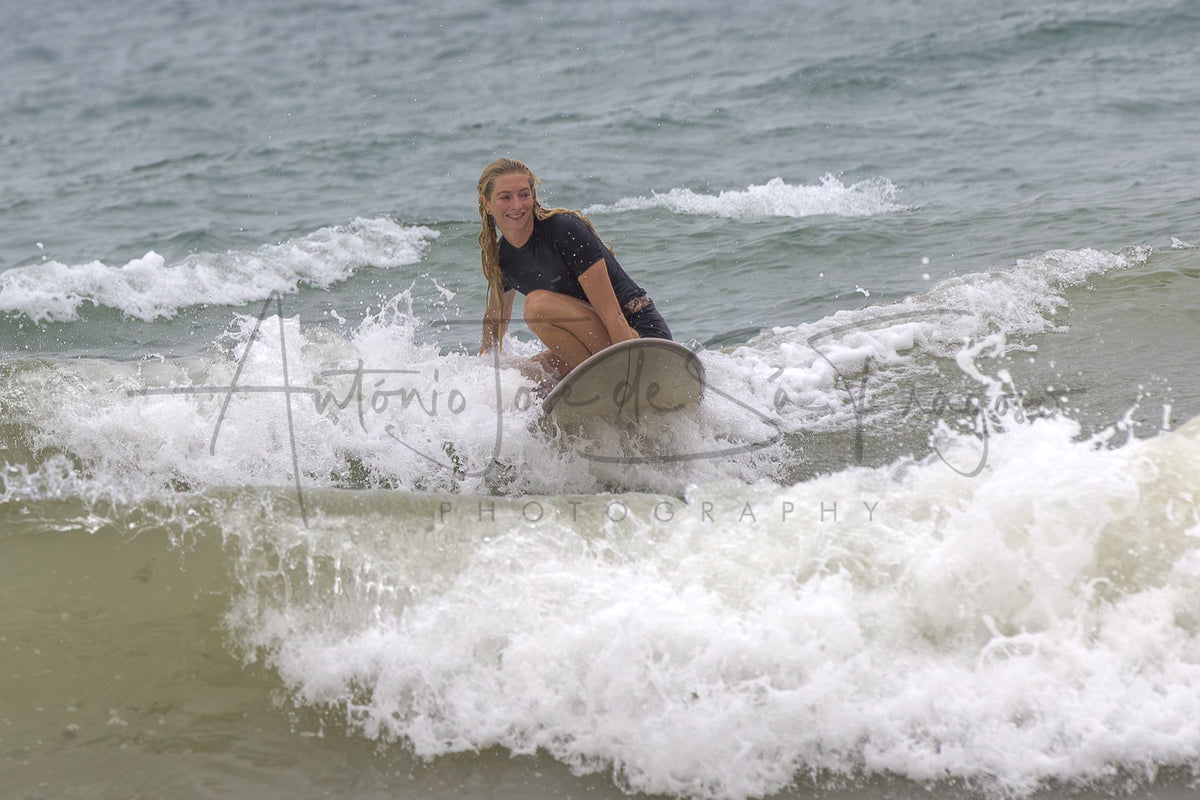 A woman in a black swimsuit crouches on a surfboard in the ocean, riding a small wave with white foam around her. A large translucent photographer watermark is overlaid across the center.